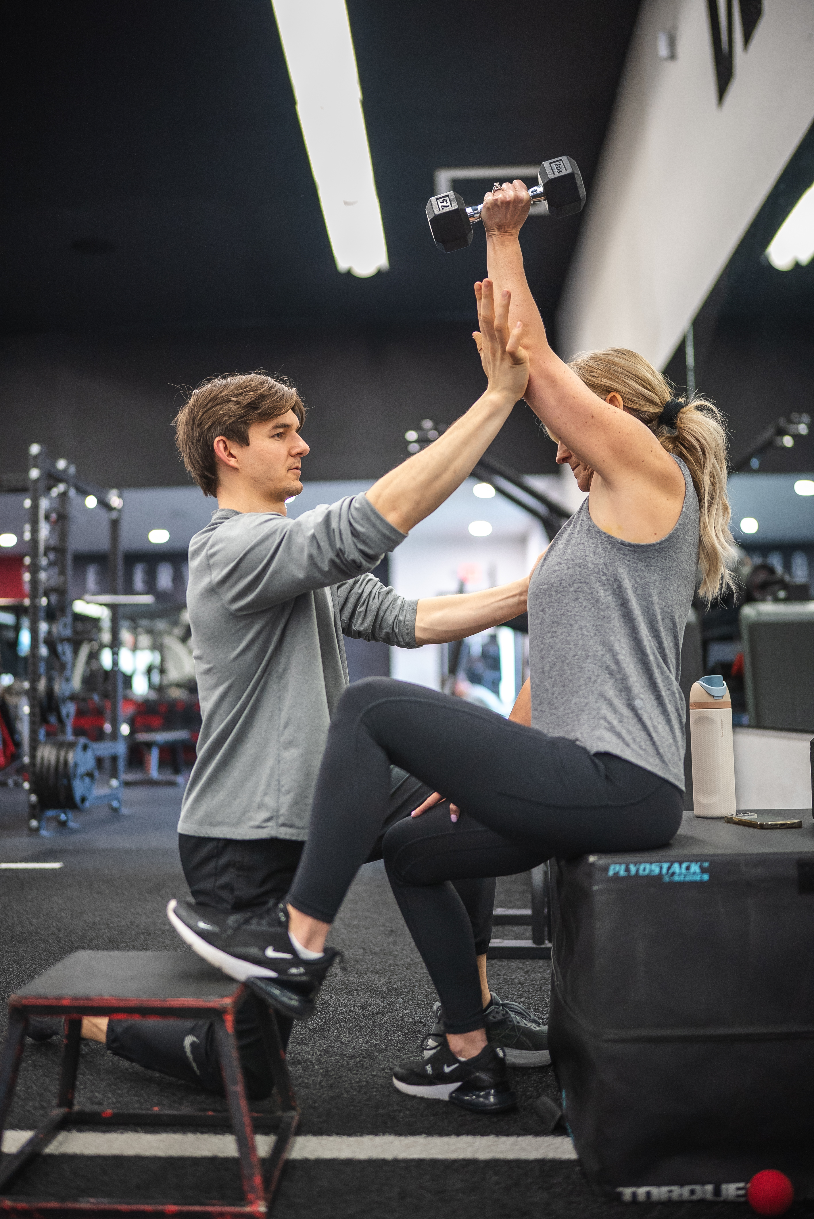 Personal trainer high-fives a smiling client as she steps onto a plyo box holding a dumbbell overhead at West Coast Strength