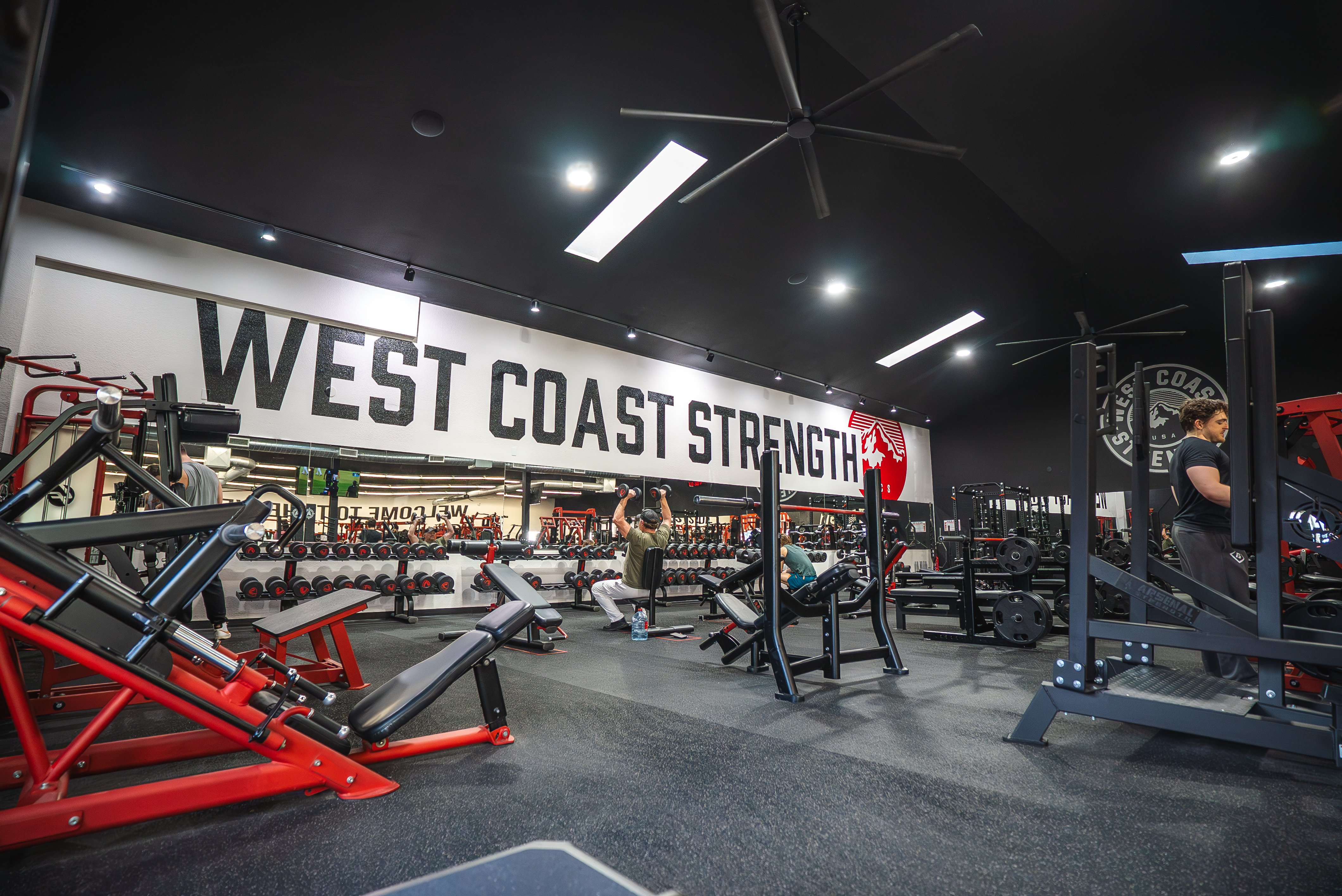 Members train among red plate-loaded machines beneath the large West Coast Strength wall lettering