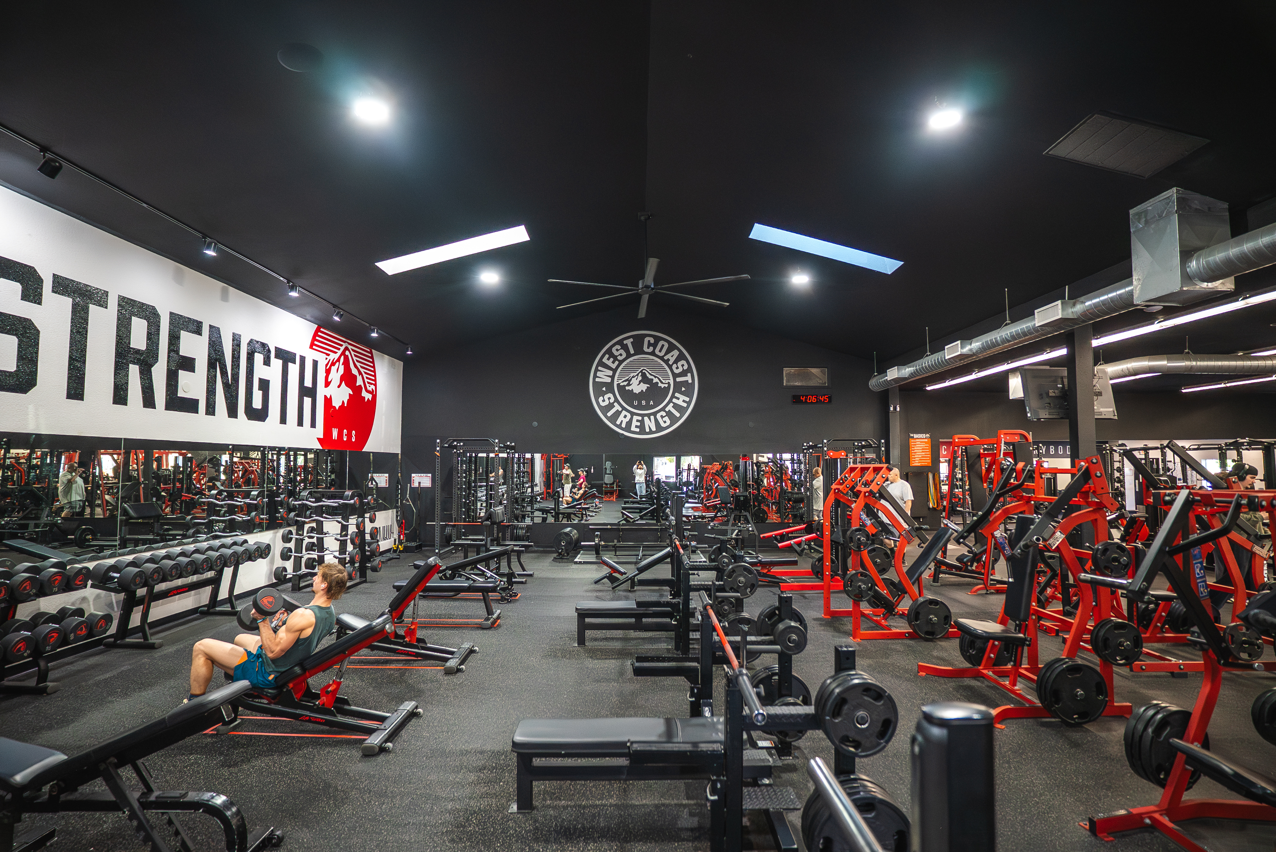 Wide view of the strength floor under a vaulted ceiling with red plate-loaded machines, benches, and the West Coast Strength mural
