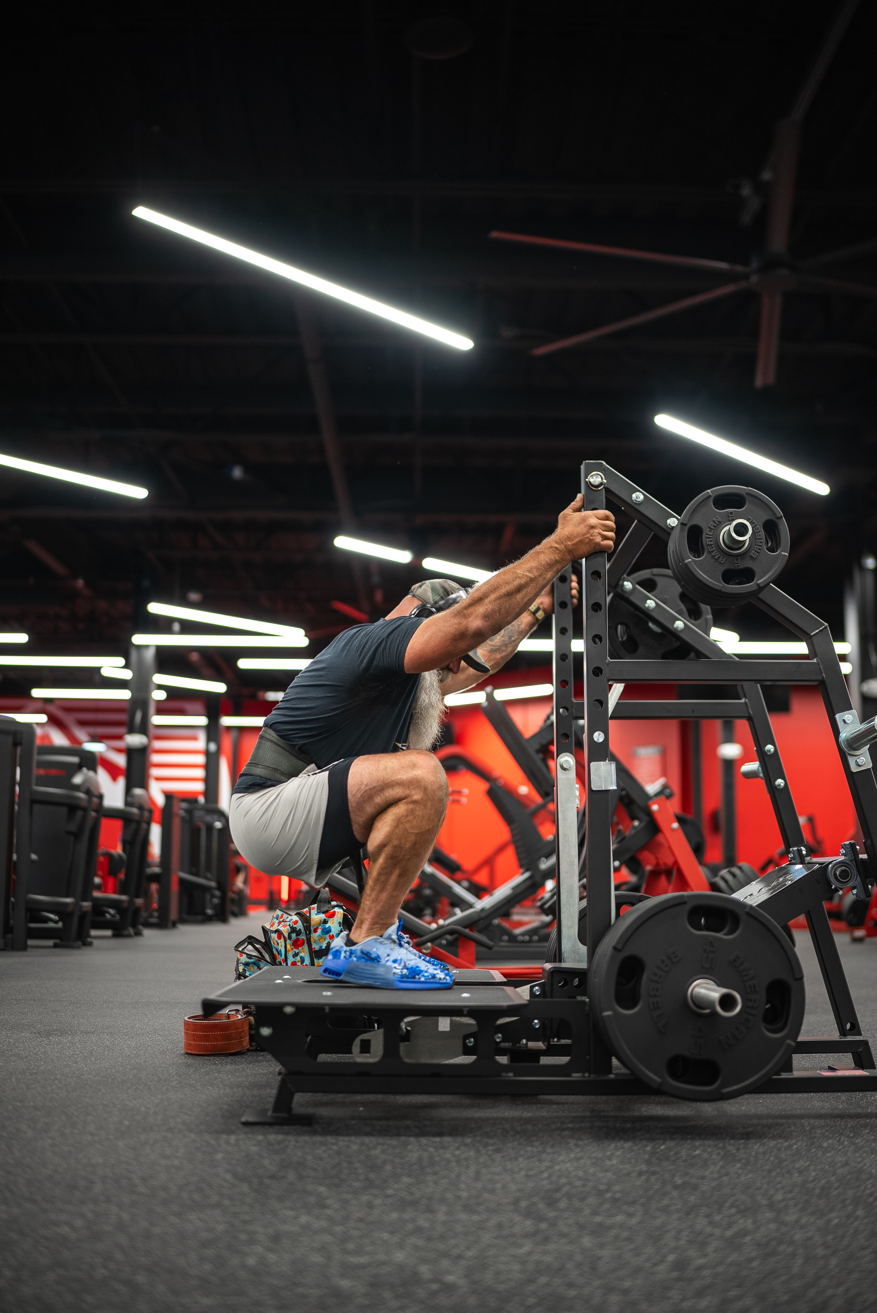 Bearded athlete performs a deep squat on a plate-loaded belt-squat machine on the gym floor
